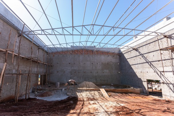 Imagem mostra a obra de um auditório, com piso ainda em chão batido. As paredes já está erguida, ainda com tijolos à vista, e a estrutura do telhado está montada, porém se cobertura. Auditório está sendo construído pela iniciativa privada em área cedida pelo Estado.