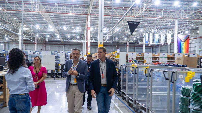 Foto em ambiente interno, em um espaço amplo, bem iluminado e com diversos carrinhos e estantes para armazenagem de produtos. Ao centro da imagem, em destaque, estão caminhando o secretário de Desenvolvimento Econômico e o representante da Amazon, lado a lado. Eles estão conversando e observando o centro de distribuição. Também aparecem outras pessoas ao fundo.