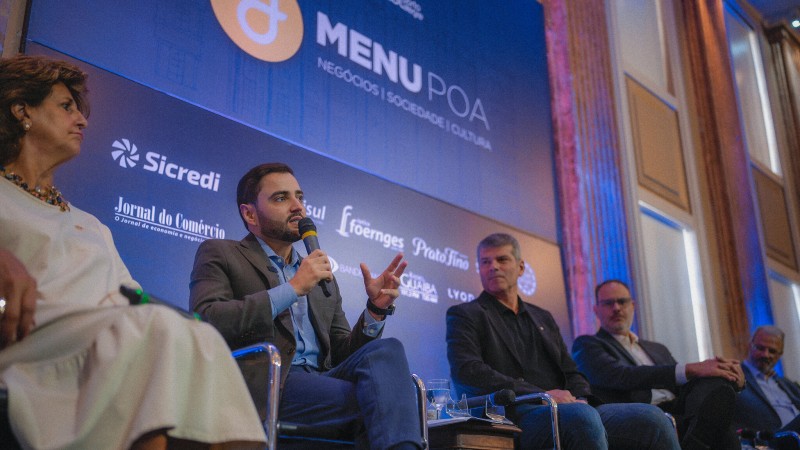 Gabriel sentado com microfone falando a uma plateia. Ao seu lado uma mulher e três homens. Eles participam de uma painel sobre o South Summit Brazil 2024. Atrás deles há um painel onde está escrito Menu Poa.