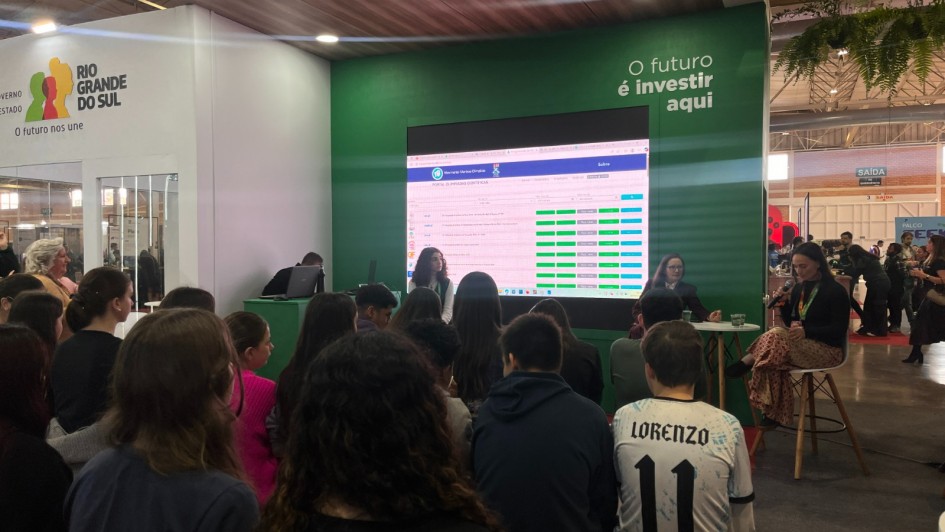 Há dezenas de crianças de costas, sentadas, assistindo uma apresentação. No palco há quatro pessoas. Dias mulheres fazem a apresentação, enquanto uma menina assiste e um homem está sentado de frente ao um notebook. Há um painel com informações atrás deles.