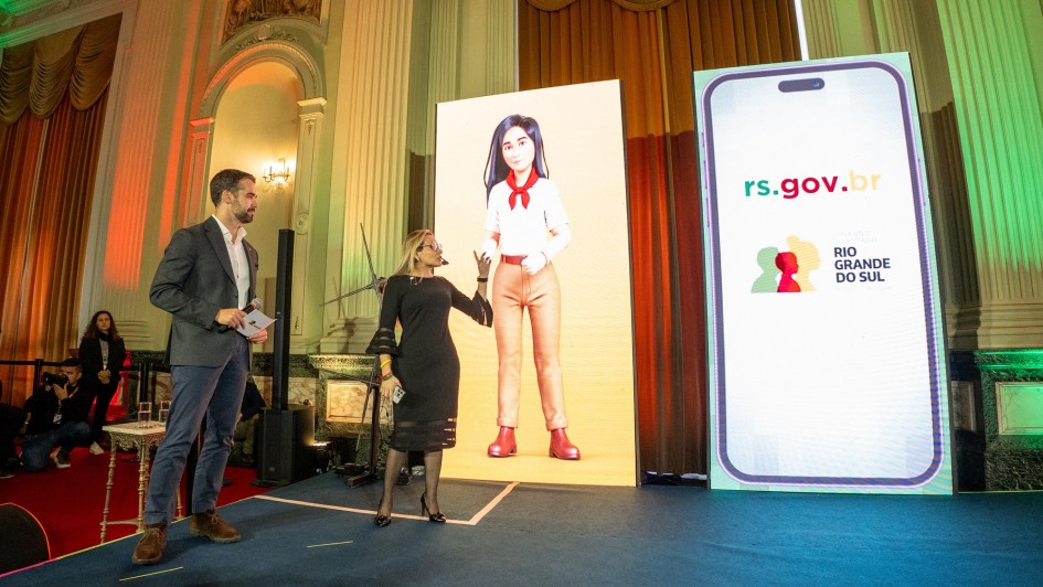 Callazans e Leite em pé sobre um palco apresentando a GurIA, inteligência artificial do rs.gov.br. Há dois cartazes grandes um deles mostrando a imagem do avatar da nova IA do Estado.