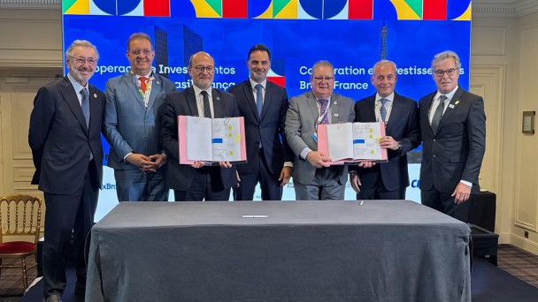 Há sete pessoas de pé posando para foto. Uma mesa com pano cinza escuro está à frente deles. Ao fundo um painel azul e com cores que lembram as bandeiras do Brasil e da França. Três seguram dois documentos abertos e assinados.