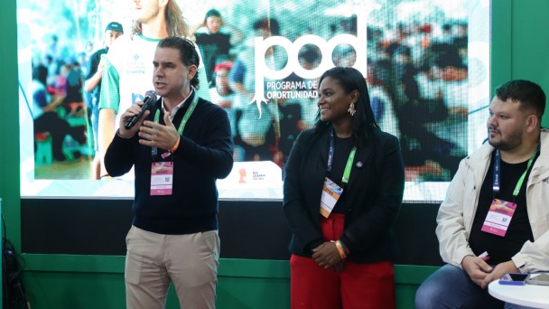 Há três pessoas em um palco. Ao fundo um painel de apresentação. Um homem está de pé e segurando um microfone. Uma mulher, ao lado dele, olha para ele. E outro homem está sentado.