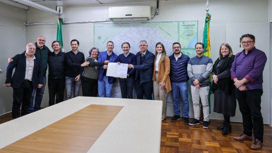 Foto posada em sala fechada. Um total de 13 pessoas está de pé, de frente para a câmera. Ao centro, o Costella exibe o documento assinado.