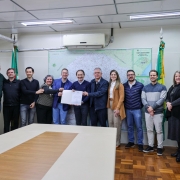 Foto posada em sala fechada. Um total de 13 pessoas está de pé, de frente para a câmera. Ao centro, o Costella exibe o documento assinado.