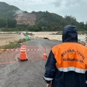Imagem mostra homem da Defesa Civil do Rio Grande do Sul olhando para água de rio que cobre ponte.