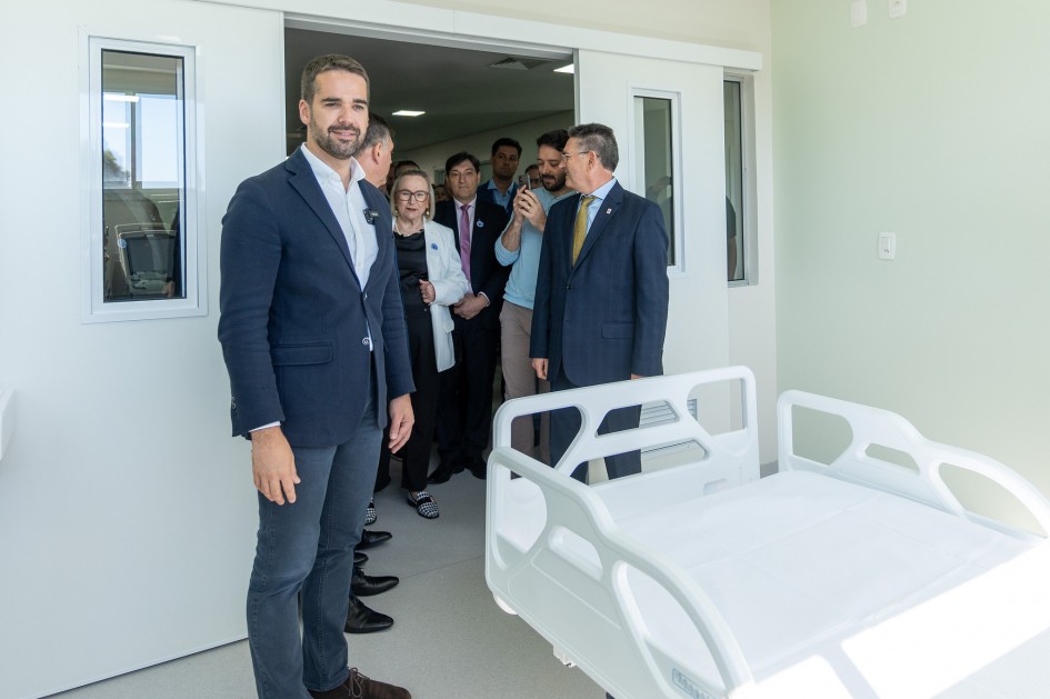 A foto mostra o Governador Eduardo Leite, um homem branco de barba escura e cabelo curto, vestindo um blazer azul-marinho, camisa branca e calça cinza escura, posando em primeiro plano, sorrindo e olhando para a câmera. Ele está em um ambiente hospitalar recém-inaugurado ou modernizado, com paredes claras e portas brancas. Atrás dele, em segundo plano, a Secretária da Saúde, Arita Bergmann, uma mulher de cabelos loiros curtos, óculos, vestindo um blazer branco, aparece conversando com outras pessoas. À direita, em primeiro plano, vê-se a parte frontal de uma cama hospitalar branca e moderna, indicando o cenário de um quarto.