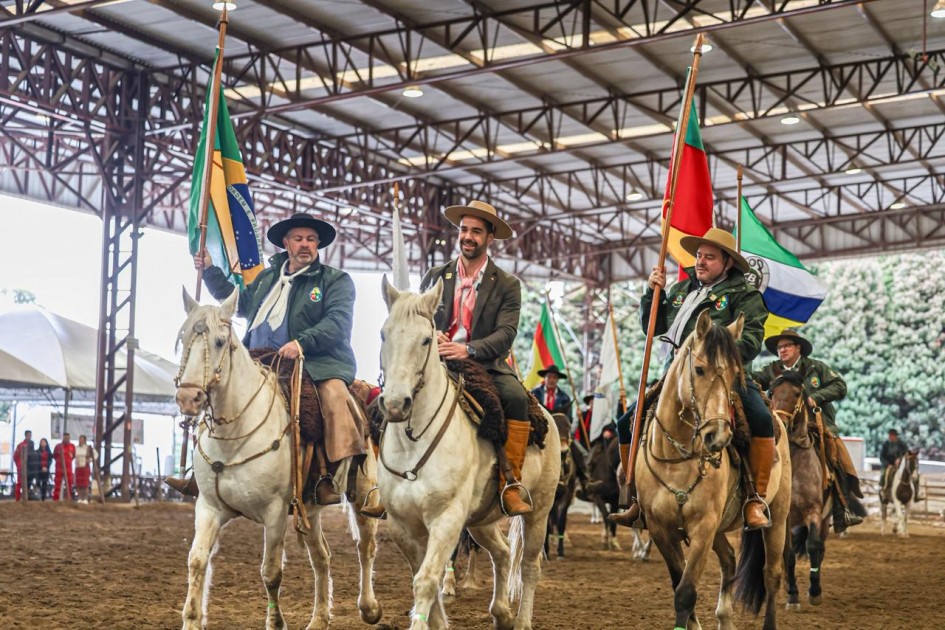 Cavaleiros em arena coberta carregam bandeiras, incluindo a do Brasil. Vestem trajes tradicionais; parece desfile ou cerimônia.