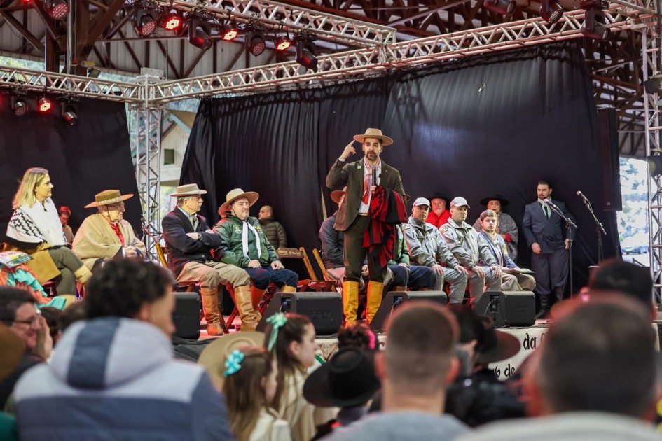 Grupo no palco com roupas tradicionais; uma pessoa fala ao microfone. Luzes vermelhas, cortina preta e público assistindo. É evento cultural.