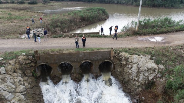 Foto de uma barragem, com &aacute;gua jorrando na metade inferior do quadro. Acima, h&aacute; cerca de 15 pessoas observando o cen&aacute;rio.