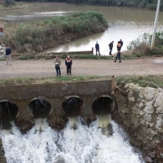 Foto de uma barragem, com &aacute;gua jorrando na metade inferior do quadro. Acima, h&aacute; cerca de 15 pessoas observando o cen&aacute;rio.