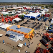 Uma fotografia a&eacute;rea colorida em plano geral e &acirc;ngulo plong&eacute;e (tomada por drone) exibe uma vis&atilde;o panor&acirc;mica e detalhada da &aacute;rea de m&aacute;quinas e implementos agr&iacute;colas da feira a c&eacute;u aberto.

**O Estande Central e a Unidade M&oacute;vel:**

* No centro-inferior da imagem, destaca-se uma edifica&ccedil;&atilde;o t&eacute;rrea retangular com paredes laterais laranjas e teto de zinco cinza, com um grande bal&atilde;o infl&aacute;vel esf&eacute;rico laranja fixado no topo.
* Logo ao lado da estrutura, encontra-se estacionada a unidade m&oacute;vel ba&uacute; com a inscri&ccedil;&atilde;o **"CARRETAS DO SABER"** em destaque na lateral azul e branca, com toldos estendidos para recep&ccedil;&atilde;o.
* Ao redor desse estande, circulam pedestres e visitantes por ruas de terra batida e cascalho claro, entre pequenas tendas de descanso com guarda-s&oacute;is brancos.

**Maquin&aacute;rios e Estandes ao Redor:**

* &Agrave; direita e na parte inferior, concentra-se uma expressiva exposi&ccedil;&atilde;o de pesados maquin&aacute;rios agr&iacute;colas em tons vivos: grandes pulverizadores autopropelidos e colheitadeiras vermelhas e laranjas, al&eacute;m de plantadeiras e tratores azuis e amarelos alinhados em fileiras.
* &Agrave; esquerda e ao centro, estendem-se dezenas de estandes comerciais com tendas e pavilh&otilde;es modulares brancos de coberturas pontiagudas ou planas, exibindo implementos agr&iacute;colas menores, tratores, ca&ccedil;ambas e carretas vermelhas. Um dos estandes principais ao fundo traz a marca **"LS Tractor"** em um galp&atilde;o azul e branco.

**O Entorno e Linha do Horizonte:**

* Na parte superior da composi&ccedil;&atilde;o, al&eacute;m do limite das ruas da feira, estende-se um imenso bols&atilde;o de estacionamento com milhares de ve&iacute;culos alinhados.
* Ao fundo, avistam-se &aacute;reas planas de v&aacute;rzea e espelhos d'&aacute;gua com vegeta&ccedil;&atilde;o baixa, al&eacute;m de uma alta torre/chamin&eacute; vertical &agrave; direita sob a linha do horizonte.
* O c&eacute;u &eacute; limpo e azul-claro com suave bruma ao longe, iluminado plenamente pela luz do sol do meio-dia.