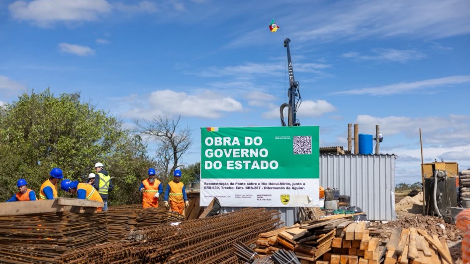 Uma fotografia colorida em plano geral e perspetiva ao n&iacute;vel dos olhos regista o canteiro de obras da reconstru&ccedil;&atilde;o de uma ponte, destacando a placa oficial do empreendimento e a equipa de trabalho.

**A Placa de Obra (ao centro):**

* No centro da composi&ccedil;&atilde;o, sobressai uma grande placa retangular com topo verde-escuro e base branca.
* No topo verde, l&ecirc;-se em letras garrafais brancas mai&uacute;sculas: **"OBRA DO GOVERNO DO ESTADO"**, com a bandeira do Rio Grande do Sul no canto superior esquerdo e um QR Code no canto superior direito.
* Na parte inferior branca, constam as informa&ccedil;&otilde;es t&eacute;cnicas e institucionais:
* **T&iacute;tulo:** *"Reconstru&ccedil;&atilde;o da Ponte sobre o Rio Ibicu&iacute;-Mirim, junto a ERS-530, Trecho: Entr. BRS-287 - Dilermando de Aguiar."*
* **Empresa e Respons&aacute;vel:** *"LAVORO ENGENHARIA LTDA | Edson Luiz da Silva CREA RS044553"*.
* **Investimento Total:** *"R$ 6.114.746,42"*.
* **Log&oacute;tipos:** A marca do *"DAER"* e do Governo do Estado na base.



**Os Oper&aacute;rios e os Materiais (em primeiro plano e &agrave; esquerda):**

* Em primeiro plano e no canto inferior esquerdo, encontram-se grandes pilhas de malhas de ferro, ferragens estruturais e t&aacute;buas de madeira empilhadas para a constru&ccedil;&atilde;o civil.
* &Agrave; esquerda, atr&aacute;s dos materiais, um grupo de oper&aacute;rios trabalha no canteiro vestindo uniformes de prote&ccedil;&atilde;o de alta visibilidade (coletes e camisas laranjas com faixas refletivas) e capacetes de seguran&ccedil;a azuis e brancos.

**As M&aacute;quinas e o Entorno (&agrave; direita e ao fundo):**

* &Agrave; direita da placa, avista-se um contentor met&aacute;lico cinzento de apoio e um dep&oacute;sito de &aacute;gua azul sobre o teto.
* Ao fundo, ergue-se a estrutura vertical de uma perfuratriz/guindaste de grande porte com uma pequena bandeira do Estado hasteada no topo.
* O c&eacute;u apresenta-se num tom azul-claro com nuvens brancas dispersas e ilumina&ccedil;&atilde;o solar direta e uniforme.
