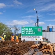 Uma fotografia colorida em plano geral e perspetiva ao n&iacute;vel dos olhos regista o canteiro de obras da reconstru&ccedil;&atilde;o de uma ponte, destacando a placa oficial do empreendimento e a equipa de trabalho.

**A Placa de Obra (ao centro):**

* No centro da composi&ccedil;&atilde;o, sobressai uma grande placa retangular com topo verde-escuro e base branca.
* No topo verde, l&ecirc;-se em letras garrafais brancas mai&uacute;sculas: **"OBRA DO GOVERNO DO ESTADO"**, com a bandeira do Rio Grande do Sul no canto superior esquerdo e um QR Code no canto superior direito.
* Na parte inferior branca, constam as informa&ccedil;&otilde;es t&eacute;cnicas e institucionais:
* **T&iacute;tulo:** *"Reconstru&ccedil;&atilde;o da Ponte sobre o Rio Ibicu&iacute;-Mirim, junto a ERS-530, Trecho: Entr. BRS-287 - Dilermando de Aguiar."*
* **Empresa e Respons&aacute;vel:** *"LAVORO ENGENHARIA LTDA | Edson Luiz da Silva CREA RS044553"*.
* **Investimento Total:** *"R$ 6.114.746,42"*.
* **Log&oacute;tipos:** A marca do *"DAER"* e do Governo do Estado na base.



**Os Oper&aacute;rios e os Materiais (em primeiro plano e &agrave; esquerda):**

* Em primeiro plano e no canto inferior esquerdo, encontram-se grandes pilhas de malhas de ferro, ferragens estruturais e t&aacute;buas de madeira empilhadas para a constru&ccedil;&atilde;o civil.
* &Agrave; esquerda, atr&aacute;s dos materiais, um grupo de oper&aacute;rios trabalha no canteiro vestindo uniformes de prote&ccedil;&atilde;o de alta visibilidade (coletes e camisas laranjas com faixas refletivas) e capacetes de seguran&ccedil;a azuis e brancos.

**As M&aacute;quinas e o Entorno (&agrave; direita e ao fundo):**

* &Agrave; direita da placa, avista-se um contentor met&aacute;lico cinzento de apoio e um dep&oacute;sito de &aacute;gua azul sobre o teto.
* Ao fundo, ergue-se a estrutura vertical de uma perfuratriz/guindaste de grande porte com uma pequena bandeira do Estado hasteada no topo.
* O c&eacute;u apresenta-se num tom azul-claro com nuvens brancas dispersas e ilumina&ccedil;&atilde;o solar direta e uniforme.