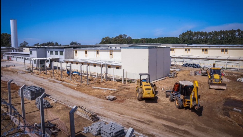 Foto mostra imagem de longe que mostra as obras na penitenci&aacute;ria de Rio Grande. Os pr&eacute;dios j&aacute; est&atilde;o pintados de cor branca. O p&aacute;tio ainda est&aacute; em terra batida tendo tr&ecirc;s retroescavadeiras trabalhando no local. 