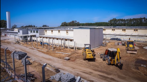 Foto mostra imagem de longe que mostra as obras na penitenci&aacute;ria de Rio Grande. Os pr&eacute;dios j&aacute; est&atilde;o pintados de cor branca. O p&aacute;tio ainda est&aacute; em terra batida tendo tr&ecirc;s retroescavadeiras trabalhando no local. 