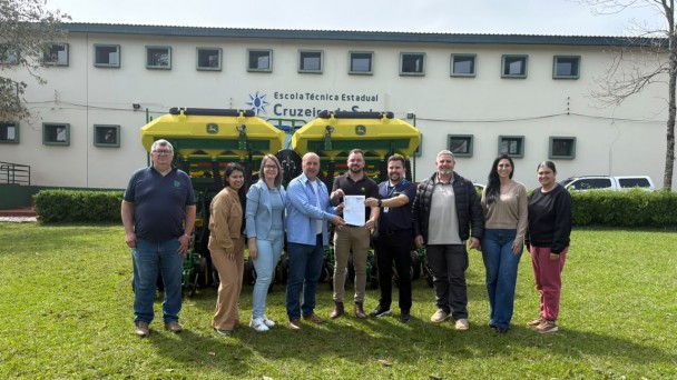Foto mostra autoridades reunidas e segurando o documento em frente &agrave; escola. Eles est&atilde;o em um gramado verde. A escola tem fachada branca. 