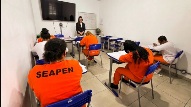 Foto mostra uma sala de aula com alunos sentados nas carteiras e uma professora em p&eacute; na frente da classe. Os alunos vestem uniformes laranjas e nas costas de um deles se l&ecirc; a sigla "Seapen".