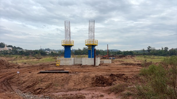 Uma fotografia colorida em plano geral e perspectiva ao n&iacute;vel dos olhos capta o canteiro de obras de constru&ccedil;&atilde;o de uma nova ponte, destacando a etapa de execu&ccedil;&atilde;o dos pilares de sustenta&ccedil;&atilde;o.

**A Infraestrutura em Constru&ccedil;&atilde;o (ao centro):**

* No centro da composi&ccedil;&atilde;o, sobressaem-se dois grandes pilares de sustenta&ccedil;&atilde;o em fase de betonagem e arma&ccedil;&atilde;o de a&ccedil;o, erguidos sobre blocos de funda&ccedil;&atilde;o maci&ccedil;os em bet&atilde;o cinzento-claro.


* A base redonda dos pilares est&aacute; envolvida por cofragens met&aacute;licas azuis, sobre as quais se encontram instaladas plataformas de trabalho e guarda-corpos de prote&ccedil;&atilde;o pintados de amarelo vibrante.


* No topo dos pilares, elevam-se as armaduras de ferro/a&ccedil;o expostas em formato cil&iacute;ndrico, aguardando as pr&oacute;ximas etapas de betonagem.



**O Canteiro de Obras e o Entorno (em primeiro plano e ao fundo):**

* Em primeiro plano e ao redor das funda&ccedil;&otilde;es, estende-se uma ampla &aacute;rea de terra batida e revolvida em tom terroso/castanho-avermelhado com marcas de pneus de m&aacute;quinas pesadas.


* Ao fundo, &agrave; direita, avistam-se m&aacute;quinas escavadoras, perfuradoras e equipamentos de constru&ccedil;&atilde;o civil em atividade no terreno.


* Ao fundo e &agrave; esquerda, observa-se uma &aacute;rea verde com vegeta&ccedil;&atilde;o densa, &aacute;rvores e ao longe a silhueta de edifica&ccedil;&otilde;es e de uma igreja rural.


* O c&eacute;u apresenta-se completamente encoberto por nuvens cinzentas e ilumina&ccedil;&atilde;o natural difusa de dia nublado.