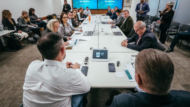 Uma fotografia colorida em plano geral e perspectiva em contra-campo (em &acirc;ngulo ligeiramente elevado) capta uma reuni&atilde;o de trabalho e alinhamento sobre a transi&ccedil;&atilde;o de governo na &aacute;rea da sa&uacute;de.

**As Autoridades e a Mesa de Reuni&atilde;o (ao centro e em primeiro plano):**

* Em primeiro plano, no centro inferior da imagem, observam-se as costas de dois participantes sentados &agrave; extremidade de uma grande mesa retangular de confer&ecirc;ncias de cor branca. O participante da esquerda veste camisa social branca e o da direita veste blazer escuro.


* Sobre o tampo da mesa, ao centro, destaca-se um tablet de controlo de videoconfer&ecirc;ncia, juntamente com telem&oacute;veis, comandos remotos, copos de &aacute;gua, blocos de notas, canetas e computadores port&aacute;teis abertos.


* Ao longo das duas extensas laterais da mesa, alinham-se cerca de uma dezena e meia de gestores, secret&aacute;rios e t&eacute;cnicos em trajes formais e esporte fino (blazers, camisas e casacos):


* &Agrave; esquerda, destaca-se a secret&aacute;ria estadual da Sa&uacute;de, Arita Bergmann, ao lado de gestoras que analisam relat&oacute;rios e documentos impressos.


* &Agrave; direita, alinham-se outros integrantes das equipas de transi&ccedil;&atilde;o consultando computadores e apontamentos.





**Os Monitores de Apresenta&ccedil;&atilde;o e o Ambiente (ao fundo):**

* Ao fundo da sala, centrados na parede principal, sobressaem dois grandes ecr&atilde;s/monitores de televis&atilde;o alinhados lado a lado transmitindo a apresenta&ccedil;&atilde;o oficial com o t&iacute;tulo: **"TRANSI&Ccedil;&Atilde;O DE GOVERNO"** sobre fundo azul-celeste.


* Ao fundo e nas extremidades da sala, outros assessores e profissionais de imprensa acompanham o encontro em p&eacute; ou dispostos em cadeiras de apoio.


* O ambiente conta com paredes em tom creme/bege claro, piso revestido a alcatifa cinzenta e ilumina&ccedil;&atilde;o embutida uniforme no teto.