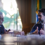 Uma fotografia colorida em plano m&eacute;dio-fechado e perspectiva ao n&iacute;vel dos olhos capta o governador Eduardo Leite em reuni&atilde;o com o prefeito de Porto Alegre, Sebasti&atilde;o Melo, no Pal&aacute;cio Piratini.

**O Governador e o Prefeito (ao centro e em primeiro plano):**

* &Agrave; direita, o governador Eduardo Leite fala de perfil diagonal voltado para a esquerda, gesticulando com ambas as m&atilde;os abertas sobre a mesa. Ele veste um fato azul-vibrante, camisa branca, gravata bege/rosa-claro e traz um pequeno pin com a bandeira do Estado na lapela do casaco.
* &Agrave; esquerda, em primeiro plano, o prefeito de Porto Alegre, Sebasti&atilde;o Melo, escuta atentamente de perfil, ligeiramente inclinado para a frente com as m&atilde;os entrela&ccedil;adas sobre a mesa. Ele possui cabelos grisalhos e usa &oacute;culos de arma&ccedil;&atilde;o escura.
* Ao seu lado, no extremo esquerdo em primeiro plano, avista-se de perfil outro gestor de fato escuro, camisa branca e gravata roxa/vinho.
* Sobre a mesa de madeira nobre em primeiro plano, veem-se copos de &aacute;gua e identificadores nominais em acr&iacute;lico desfocados pela profundidade de campo reduzida.

**O Ambiente do Sal&atilde;o (ao fundo):**

* Ao fundo, entre as figuras principais, uma grande janela iluminada revela a vegeta&ccedil;&atilde;o verde do exterior atrav&eacute;s de um vidro amplo e cortinados dourados recolhidos.
* &Agrave; direita de Eduardo Leite, avista-se parcialmente a Bandeira do Rio Grande do Sul (nas cores verde, vermelha e amarela) disposta num hasteamento interior.
* A ilumina&ccedil;&atilde;o natural proveniente da janela ao fundo cria um forte contraste de contraluz, destacando os contornos dos participantes na mesa de reuni&atilde;o.
