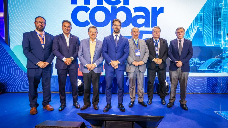 Uma fotografia colorida em plano geral e perspectiva ao n&iacute;vel dos olhos registra o governador Eduardo Leite acompanhado por autoridades e lideran&ccedil;as empresariais no palco de abertura da Mercopar, em Caxias do Sul.

**As Autoridades e Lideran&ccedil;as no Palco (ao centro e em primeiro plano):**

* No centro de um palco coberto por alcatifa azul, sete homens posam alinhados lado a lado para a fotografia oficial com as m&atilde;os entrela&ccedil;adas &agrave; frente do corpo.


* Ao centro, o governador Eduardo Leite sorri de fato azul-bic/azul-vibrante, camisa social branca e gravata azul-marinho.


* &Agrave; sua direita (&agrave; esquerda do observador), alinham-se tr&ecirc;s gestores e lideran&ccedil;as: um homem de fato azul-marinho de &oacute;culos e gravata salm&atilde;o; um gestor de fato azul-escuro sem gravata; e um homem de blazer azul-claro com camisola amarela por baixo de camisa social.


* &Agrave; sua esquerda (&agrave; direita do observador), encontram-se outros tr&ecirc;s representantes: um homem de fato cinzento-claro com gravata verde; um executivo de fato escuro com crach&aacute; oficial do evento; e um homem de fato azul-escuro, camisa clara e gravata escura na extremidade direita.


* Em primeiro plano, no piso do palco &agrave; frente do grupo, observam-se monitores de retorno pretos e caixas de som viradas para os palestrantes.



**O Painel do Evento e a Cenografia (ao fundo):**

* Ao fundo, destaca-se um grande ecr&atilde; de LED azul e branco com o log&oacute;tipo oficial do evento em letras garrafais mai&uacute;sculas: **"mercopar 33&ordf;"** (ou indica&ccedil;&atilde;o da edi&ccedil;&atilde;o) e o cabe&ccedil;alho **"FEIRA DE INOVA&Ccedil;&Atilde;O INDUSTRIAL"**.


* &Agrave; direita no painel, veem-se ilustra&ccedil;&otilde;es e grafismos de pe&ccedil;as industriais e tecnologia de precis&atilde;o sob ilumina&ccedil;&atilde;o azulada de palco.
