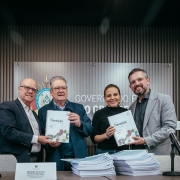 Uma fotografia colorida em plano m&eacute;dio-geral e perspectiva ao n&iacute;vel dos olhos capta quatro autoridades estaduais apresentando os Livros de Transi&ccedil;&atilde;o oficializados para subsidiar a pr&oacute;xima gest&atilde;o governamental.

**As Autoridades e os Livros de Transi&ccedil;&atilde;o (ao centro e em primeiro plano):**

* No centro da composi&ccedil;&atilde;o, quatro gestores posam alinhados lado a lado atr&aacute;s de uma mesa de confer&ecirc;ncias:
* &Agrave; esquerda, um homem de cabelos grisalhos curtos e &oacute;culos veste um blazer azul-escuro sobre camisa clara e junta-se ao colega ao lado para segurar um exemplar encadernado.
* Ao seu lado, outro gestor de cabelos grisalhos e &oacute;culos segura com ambas as m&atilde;os o livro de capa clara com espiral branca onde se l&ecirc; em destaque a palavra **"Transi&ccedil;&atilde;o"** acompanhada de grafismos geom&eacute;tricos coloridos.
* &Agrave; sua direita, a secret&aacute;ria de Planejamento, Governan&ccedil;a e Gest&atilde;o, Danielle Calazans, sorri vestindo blusa escura de mangas compridas e segura outro exemplar do Livro de Transi&ccedil;&atilde;o com o mesmo grafismo institucional.
* Na extremidade direita, um gestor de barba e cabelo curto veste blazer cinzento-claro sobre camisa escura, segurando o exemplar ao lado da secret&aacute;ria.


* Em primeiro plano, sobre o tampo da mesa, empilham-se dezenas de relat&oacute;rios e documentos oficiais encadernados em espiral branca.
* Nas extremidades da mesa, destacam-se dois microfones com base de mesa e identificadores em acr&iacute;lico com placas nominais.

**O Painel do Fundo e o Ambiente (ao fundo):**

* Ao fundo, a parede de fundo &eacute; revestida por ripas verticais de madeira em tom castanho-m&eacute;dio.
* No centro da parede, sobressai um painel acr&iacute;lico transparente com o bras&atilde;o oficial do Estado do Rio Grande do Sul e as letras mai&uacute;sculas em relevo met&aacute;lico de parte do texto institucional: **"GOVERNO DO ESTADO"**.
* A ilumina&ccedil;&atilde;o no interior do espa&ccedil;o &eacute; limpa e focada sobre as autoridades, destacando o ato solene de entrega dos documentos.